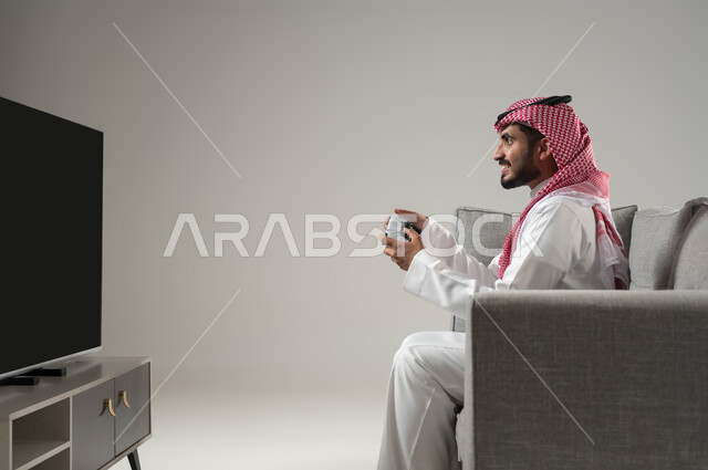 مفهوم الفوز والخسارة, العاب البلايستيشن الترفيهية والممتعة, استخدام اجهزة تقنية حديثة متطورة, قضاء وقت ممتع بألعاب الفيديو, بورتيه لشاب عربي سعودي خليجي يرتدي الثوب التقليدي والشماغ يجلس على اريكة مريحة يمسك بيده عصا التحكم, خلفية رمادية