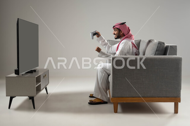 Fun and entertainment in video games, joy and happiness in achieving victory, spending a good time, interacting and merging with PlayStation games, advanced and entertaining modern technologies, a portrait of a young Saudi Arabian Gulf man wearing the traditional dress and shemagh, holding a joystick in his hand, gray background.