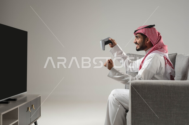 الفرح والسعادة بتحقيق الفوز, تعابير التركيز والاندماج في اللعب, قضاء وقت ممتع, التفاعل مع العاب البلايستيشن, تقنيات حديثة متطورة ومسلية, بورتريه لشاب عربي سعودي خليجي يرتدي الثوب التقليدي والشماغ يمسك بيده عصا التحكم, خلفية رمادية