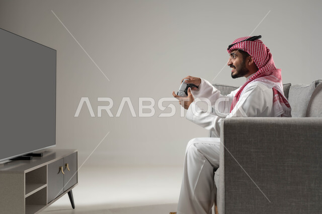 قضاء وقت ممتع, التفاعل والاندماج مع العاب البلايستيشن, تقنيات حديثة متطورة ومسلية, بورتريه لشاب عربي سعودي خليجي يرتدي الثوب التقليدي والشماغ يمسك بيده عصا التحكم, تعابير التركيز في اللعب, خلفية رمادية