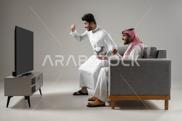 Jumping with joy when the favorite team wins, expressions of enthusiasm and victory, sports matches and games, portrait of two Saudi Gulf Arab men, one of them holding a football in his hand, passion for recreational activities, spending a good time with friends, gray background