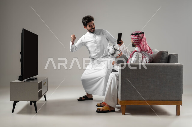 تعابير الفوز والانتصار, النظر إلى كاميرا الموبايل بإيماءات السعادة والحماس، استخدام التكنولوجيا الحديثة، بورتريه برجلان عربيان سعوديان خليجيان يجلسان على كنبة مريحة يأخذان صور تذكارية، خلفية رمادية