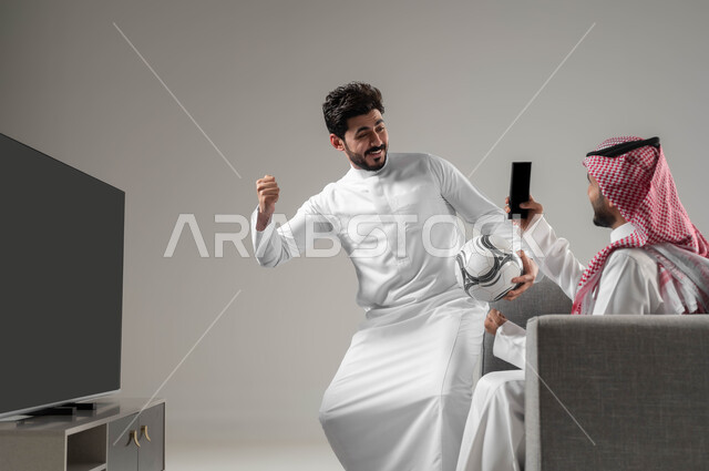 النظر إلى كاميرا الموبايل بإيماءات السعادة والحماس، استخدام التكنولوجيا الحديثة، بورتريه برجلان عربيان سعوديان خليجيان يجلسان على كنبة مريحة يأخذان صور تذكارية, تعابير الفوز والانتصار، خلفية رمادية