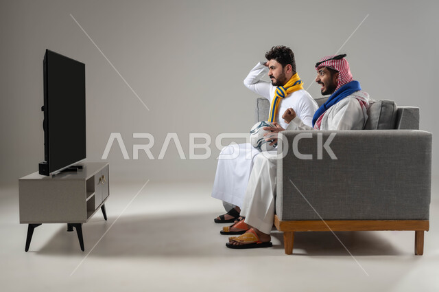 المنافسة القوية بين الفرق الرياضية, تعابير التحدي والخسارة، انشطة ترفيهية في وقت الفراغ, بورتريه من الجانب لرجلان عربيان خليجيان سعوديان مبتسمان يرتديان أوشحة تشجيعية لفرق كرة القدم، قضاء وقت ممتع مع الأصدقاء, مشاهدة التلفاز مع الزملاء، خلفية رمادية