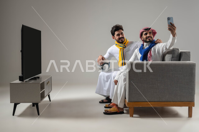 Live video broadcast, use of modern electronic devices, close-up portrait of two smiling Saudi Gulf Arab men wearing football team scarves, taking a souvenir photo using a mobile phone, selfies, front camera, spending a good time with friends, gray background