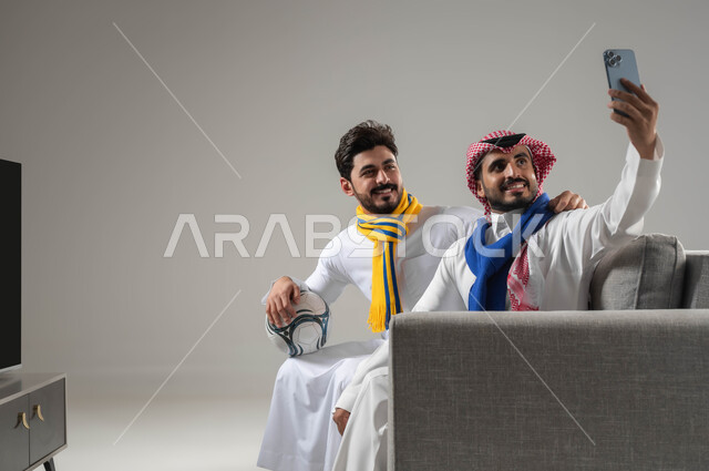 صور تذكارية، بورتريه مقرب لرجلان عربيان خليجيان سعوديان مبتسمان يرتديان أوشحة تشجيعية لفرق كرة القدم يقومان بالتقاط صورة تذكارية من خلال الهاتف المحمول، كاميرا امامية، بث فيديو مباشر، استخدام أجهزة الكترونية حديثة، قضاء وقت ممتع مع الأصدقاء، خلفية رمادية