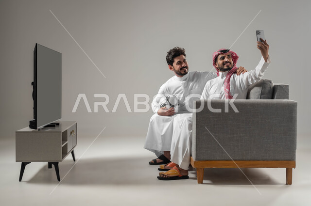 الاستمتاع بأخذ صور سيلفي، استخدام أجهزة الكترونية حديثة، بورتريه لرجلان عربيان خليجيان سعوديان مبتسمان يقومان بالتقاط صورة تذكارية من خلال الهاتف المحمول، توثيق اللحظات السعيدة، بث فيديو مباشر، قضاء وقت ممتع مع الأصدقاء، خلفية رمادية