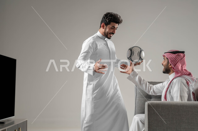 قضاء أوقات الفراغ بمشاهدة التلفاز مع الأصدقاء، بورتريه مقرب من الجانب لرجلان عربيان سعوديان خليجيان يحتفلان بفوز فريقهم المفضل ، متابعة المباريات والالعاب الرياضية، ايماءات الشغف والسعادة والحماس، خلفية رمادية