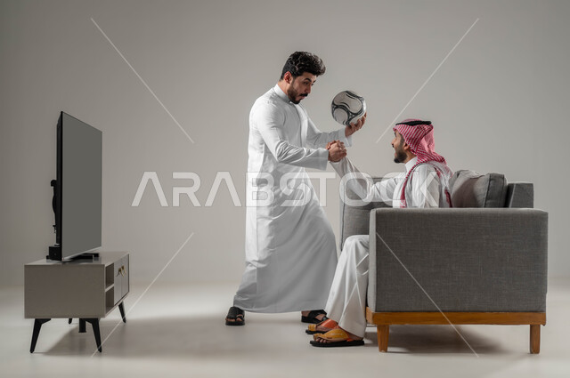 أنشطة ترفيهية منزلية، مفهوم التحدي والمنافسة، قضاء أوقات ممتعة مع الأصدقاء، بورتريه من الجانب لرجلان عربيان خليجيان سعوديان يقومان بمتابعة المباريات والألعاب الرياضية، النظر بإيماءات الشغف والحماس، خلفية رمادية