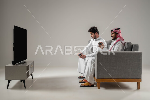 ألعاب البلايستيشين الممتعة، قضاء أوقات الفراغ بالألعاب الترفيهية مع الأصدقاء،  بورتريه من الجانب لرجلان عربيان سعوديان خليجيان يمسكان عصا التحكم يستمتعان باللعب الجماعي، تقنيات حديثة مسلية , مفهوم التحدي والمنافسة، النظر بإيماءات الحماس للفوز، خلفية رمادية