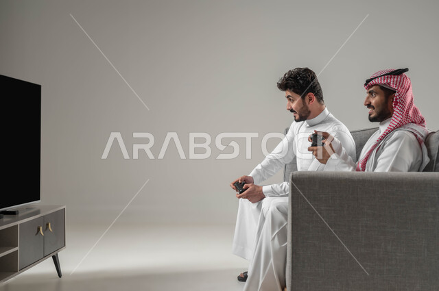 Entertaining modern technologies, the concept of challenge and competition, spending leisure time playing entertainment PlayStation games, a close-up portrait from the side of two Saudi Gulf Arab men holding a joystick enjoying group play, looking with happy gestures for victory, gray background
