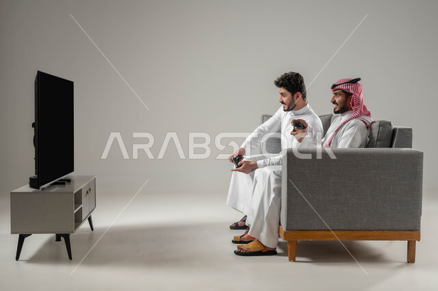 مفهوم التحدي والمنافسة، قضاء أوقات الفراغ باللعب بألعاب البلايستيشن الترفيهية، بورتريه من الجانب لرجلان عربيان خليجيان سعوديان يجلسان على أريكة يحملان عصا التحكم، النظر بإيماءات السعادة للفوز، خلفية رمادية