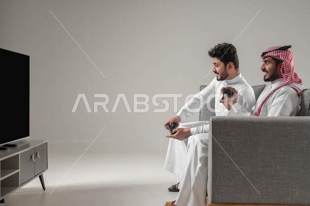 قضاء وقت الفراغ بألعاب الفيديو مع الاصدقاء, الالعاب الالكترونية الترفيهية والممتعة, بورتريه مقرب لرجلان عربيان سعوديان خليجيان يمسكان عصا التحكم يستمتعان اللعب الجماعي, تقنيات واجهزة متطورة مسلية, ايماءات الفرح والسرور، خلفية رمادية