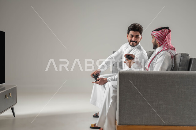 تعابير التحدي والمواجهة, الاستمتاع باللعب الجماعي في المنزل, الالعاب الالكترونية الترفيهية والممتعة, تقنيات واجهزة متطورة مسلية, بورتريه لرجلان عربيان سعوديان خليجيان يمسكان عصا التحكم, قضاء وقت الفراغ بألعاب الفيديو مع الاصدقاء, خلفية رمادية