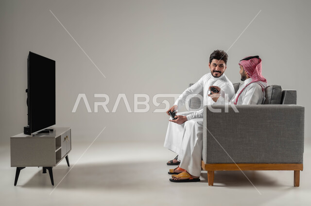 الاستمتاع باللعب الجماعي في المنزل, الالعاب الالكترونية الترفيهية والممتعة, تقنيات واجهزة متطورة مسلية, تعابير التحدي والمواجهة, بورتريه لرجلان عربيان سعوديان خليجيان يمسكان عصا التحكم, قضاء وقت الفراغ بألعاب الفيديو مع الاصدقاء, خلفية رمادية
