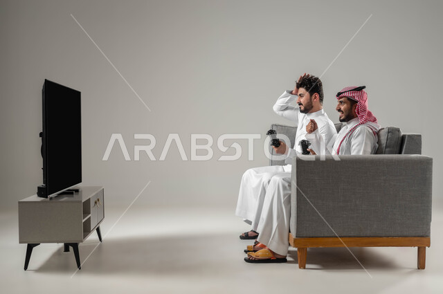 Entertaining and fun electronic games, advanced entertaining technologies and devices, expressions of loss and sadness, portrait of two Saudi-Gulf Arab men holding a joystick, spending free time playing video games with friends, gray background