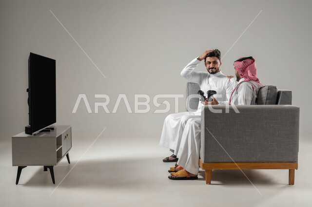 Entertaining advanced technologies and devices, expressions of loss and sadness, recreational PlayStation games, portrait of two Saudi-Gulf Arab men holding a joystick, spending free time playing video games with friends, gray background