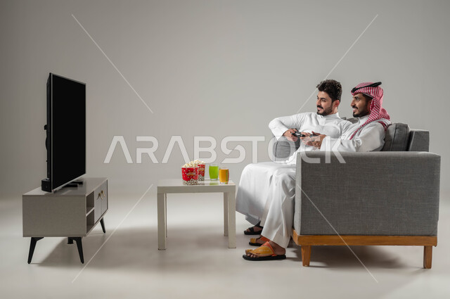 العاب البلاستيشن الترفيهية, قضاء وقت الفراغ بألعاب الفيديو مع الاصدقاء, بورتريه لرجلان عربيان سعوديان خليجيان يمسكان عصا التحكم, تناول الوجبات الخفيفة, تقنيات واجهزة متطورة مسلية, الاستمتاع باللعب الجماعي في المنزل, خلفية رمادية
