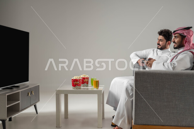 Spending free time playing video games with friends, recreational PlayStation games, a portrait of two Saudi Arabian Gulf men holding a joystick, eating snacks, advanced entertaining technologies and devices, enjoying group play at home, gray background