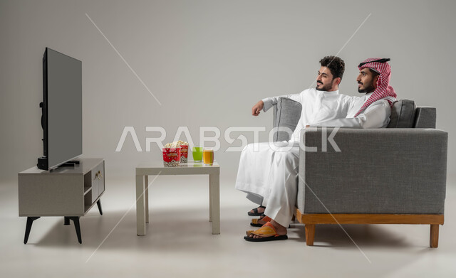 تعابير التركيز والاندماج, الاستمتاع بمشاهدة التلفاز مع الاصدقاء, مفهوم العلاقة لقوية والترابط بين الزملاء, تعابير التسلية والمرح, بورتريه لرجلان عربيان سعوديان خليجيان, نشاط منزلي ترفيهي جماعي, قضاء اوقات ممتعة ومسلية, تناول الاطعمة الخفيفة, خلفية رمادية