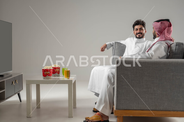مفهوم العلاقة لقوية والترابط بين الزملاء, الاستمتاع بتبادل الاحاديث بين الصديقين, تعابير التسلية والمرح, بورتريه لرجلان عربيان سعوديان خليجيان, نشاط منزلي ترفيهي جماعي, قضاء اوقات ممتعة ومسلية مع الاصدقاء, تناول الاطعمة الخفيفة, خلفية رمادية