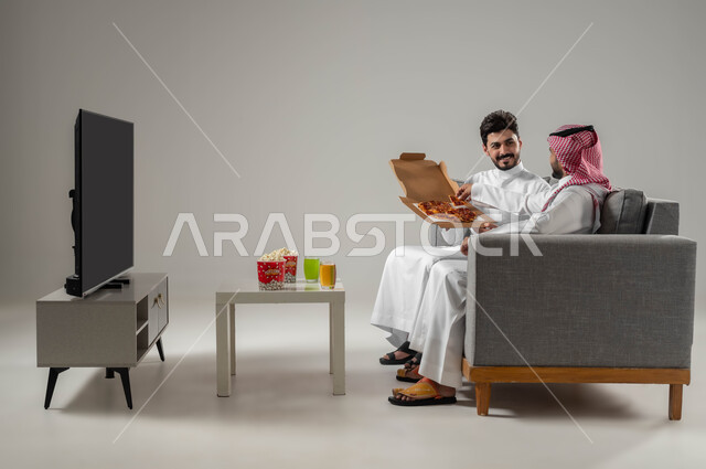 Expressions of amusement and fun, enjoying eating pizza and snacks, a portrait of two Saudi Gulf Arab men, a group recreational home activity, spending fun and entertaining times with friends, the concept of a strong relationship and interconnectedness between colleagues, gray background