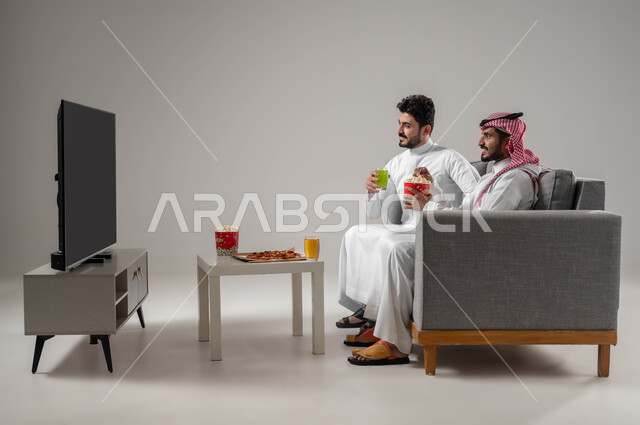 تعابير التركيز والاندماج في الفيلم, قضاء اوقات ممتعة ومسلية مع الاصدقاء, بورتريه لرجلان عربيان سعوديان خليجيان, الاستمتاع بتناول الفشار والاطعمة الخفيفة اثناء مشاهدة التلفاز, نشاط منزلي ترفيهي جماعي, خلفية رمادية
