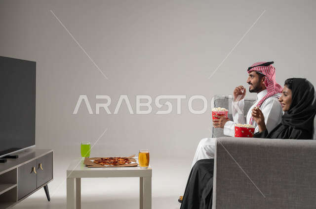 A happy family atmosphere, focus and involvement in watching, enjoying spending leisure time with the family, portrait of a Saudi-Gulf Arab couple sitting in the living room, watching TV with gestures of amusement and joy, eating light foods, gray background