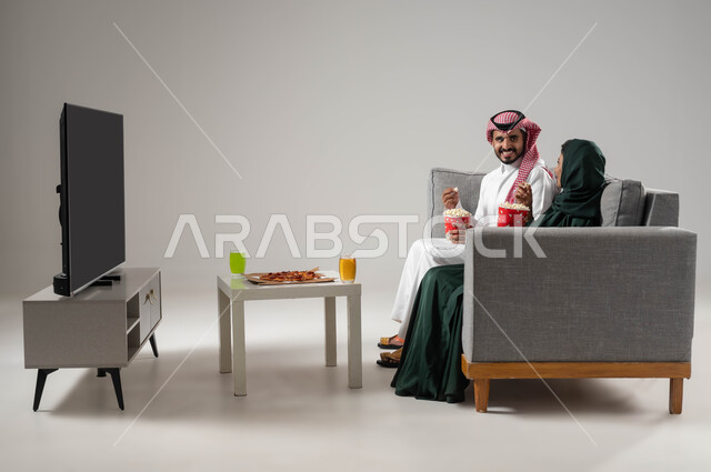 قضاء اوقات ممتعة ومسلية, مفهوم الترابط والتفاهم بين الزوجين, الاستمتاع بقضاء أوقات ترفيهية مع العائلة, بورتريه لزوجان عربيان سعوديان خليجيان يجلسان في غرفة المعيشة, مشاهدة التلفاز بإيماءات المحبة والفرح, تناول الأطعمة الخفيفة, خلفية رمادية