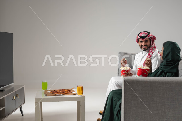 مفهوم الترابط والتفاهم بين الزوجين, الاستمتاع بقضاء أوقات ترفيهية مع العائلة, قضاء اوقات ممتعة ومسلية, بورتريه لزوجان عربيان سعوديان خليجيان يجلسان في غرفة المعيشة, مشاهدة التلفاز بإيماءات المحبة والفرح, تناول الأطعمة الخفيفة, خلفية رمادية