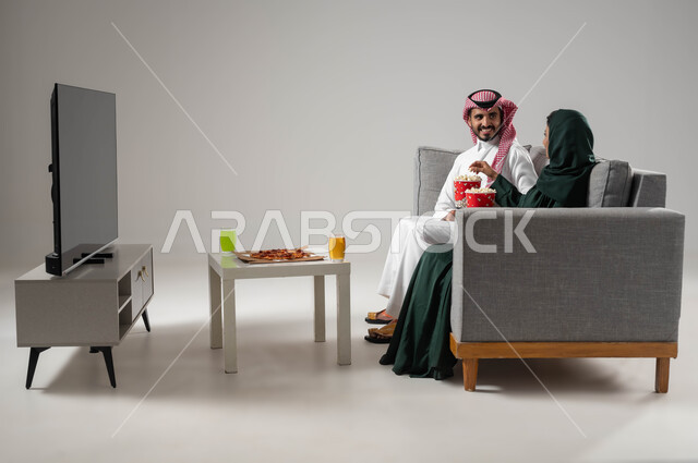 الاستمتاع بقضاء أوقات ترفيهية مع العائلة, قضاء اوقات ممتعة ومسلية, بورتريه لزوجان عربيان سعوديان خليجيان يجلسان في غرفة المعيشة, مشاهدة التلفاز بإيماءات المحبة والفرح, تناول الأطعمة الخفيفة, خلفية رمادية