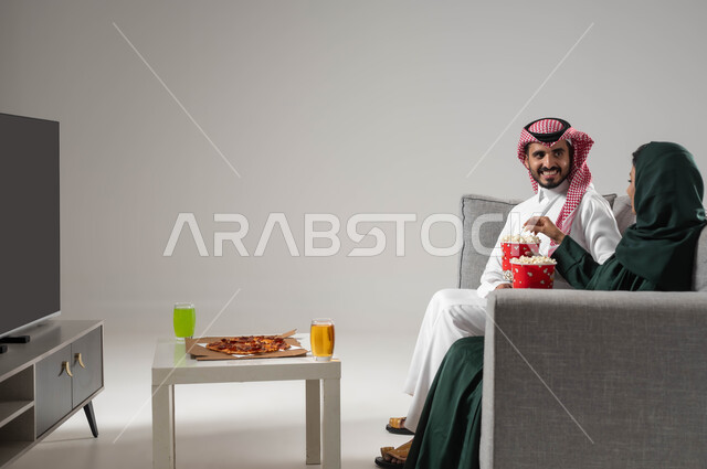 تناول وجبات خفيفة، استخدام التكنولوجيا في الترفيه، مشاهدة التلفاز والاستمتاع بقضاء الوقت مع العائلة، مفهوم الترابط و التفاهم الأسري، بورتريه من الجانب لإمرأة ورجل عربيين سعوديين خليجيين يجلسان على اريكة مريحة يحملان دلو الفشار، صورة لكامل الجسم، خلفية رمادية