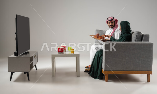 استخدام التكنولوجيا في الترفيه، تناول وجبة خفيفة ومشاهدة التلفاز والاستمتاع بقضاء الوقت مع العائلة، مفهوم الترابط و التفاهم الأسري، بورتريه من الجانب لإمرأة ورجل عربيين سعوديين خليجيين يجلسان على اريكة مريحة يحملان علبة بيتزا، صورة لكامل الجسم، خلفية رمادية