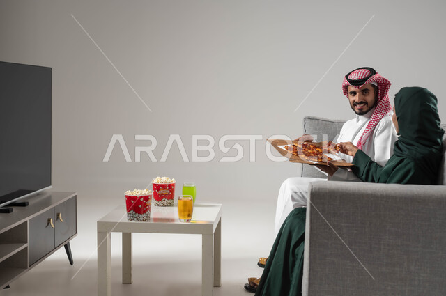 تناول وجبة خفيفة ومشاهدة التلفاز والاستمتاع بقضاء الوقت مع العائلة، مفهوم الترابط و التفاهم الأسري، بورتريه من الجانب لإمرأة ورجل عربيين سعوديين خليجيين يجلسان على اريكة مريحة يحملان علبة بيتزا، استخدام التكنولوجيا بالترفيه، صورة لكامل الجسم، خلفية رمادية