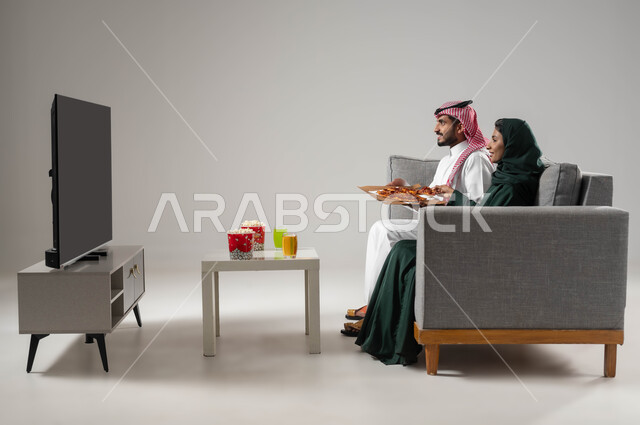 مفهوم الترابط و التفاهم الأسري، بورتريه من الجانب لإمرأة ورجل عربيين سعوديين خليجيين يجلسان على اريكة مريحة يحملان علبة بيتزا، مشاهدة التلفاز والاستمتاع بقضاء الوقت مع العائلة، تناول وجبة خفيفة، استخدام التكنولوجيا بالترفيه، صورة لكامل الجسم، خلفية رمادية