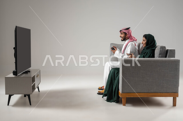 اللعب بألعاب البلايستيشن الترفيهية، قضاء أوقات عائلية ممتعة، استخدام عصا التحكم، إيماءات السعادة للفوز، مفهوم التحدي والمنافسة، تقنيات حديثة مسلية, بورتريه من الجانب لإمرأة ورجل عربيان سعوديان خليجيان محجبان يجلسان على اريكة مريحة، صورة لكامل الجسم، خلفية رمادية