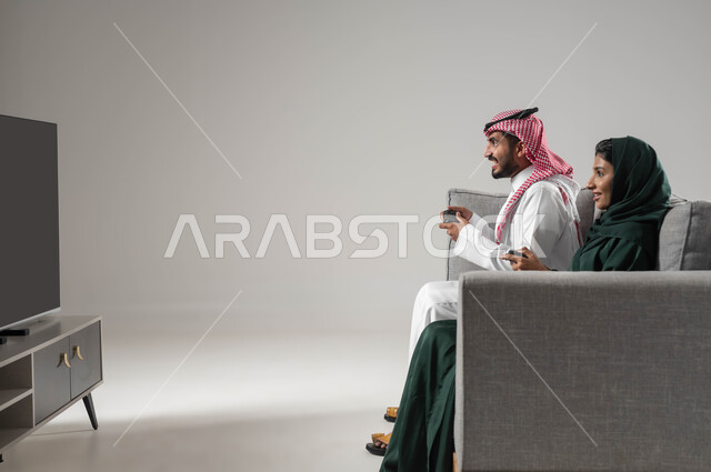 قضاء أوقات عائلية ممتعة، استخدام عصا التحكم، اللعب بألعاب البلايستيشن الترفيهية،  إيماءات السعادة للفوز، مفهوم التحدي والمنافسة، تقنيات حديثة مسلية, بورتريه من الجانب لإمرأة ورجل عربيان سعوديان خليجيان محجبان يجلسان على اريكة مريحة، صورة لكامل الجسم، خلفية رمادية