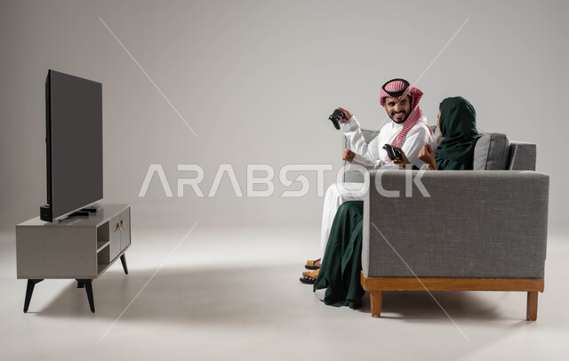 استخدام عصا التحكم، قضاء أوقات الفراغ باللعب بألعاب البلايستيشن الترفيهية،  إيماءات السعادة للفوز، مفهوم التحدي والمنافسة، تقنيات حديثة مسلية, بورتريه من الجانب لإمرأة ورجل عربيان سعوديان خليجيان محجبان يجلسان على اريكة مريحة، صورة لكامل الجسم، خلفية رمادية