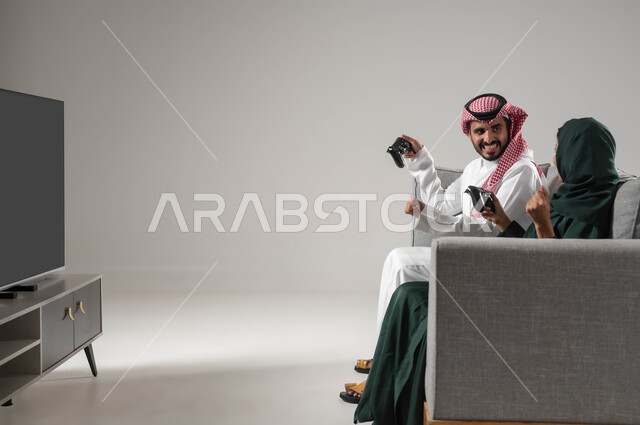 إيماءات السعادة للفوز، استخدام عصا التحكم، قضاء أوقات الفراغ باللعب بألعاب البلايستيشن الترفيهية، مفهوم التحدي والمنافسة، تقنيات حديثة مسلية, بورتريه من الجانب لإمرأة ورجل عربيان سعوديان خليجيان محجبان يجلسان على اريكة مريحة، صورة لكامل الجسم، خلفية رمادية