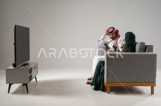 إيماءات الحزن للخسارة، استخدام عصا التحكم، قضاء أوقات الفراغ باللعب بألعاب البلايستيشن الترفيهية، مفهوم التحدي والمنافسة، تقنيات حديثة مسلية, بورتريه من الجانب لإمرأة ورجل عربيان سعوديان خليجيان محجبان يجلسان على اريكة مريحة، صورة لكامل الجسم، خلفية رمادية