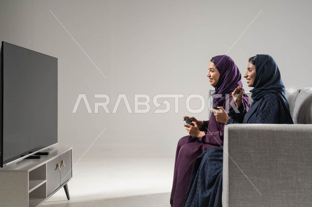استخدام عصا التحكم، قضاء أوقات الفراغ باللعب بألعاب البلايستيشن الترفيهية، مفهوم التحدي والمنافسة، النظر بإيماءات السعادة للفوز، تقنيات حديثة مسلية, بورتريه من الجانب لإمرأتان عربيتان سعوديتان خليجيتان محجبتان تجلسان على اريكة، صورة لكامل الجسم، خلفية رمادية