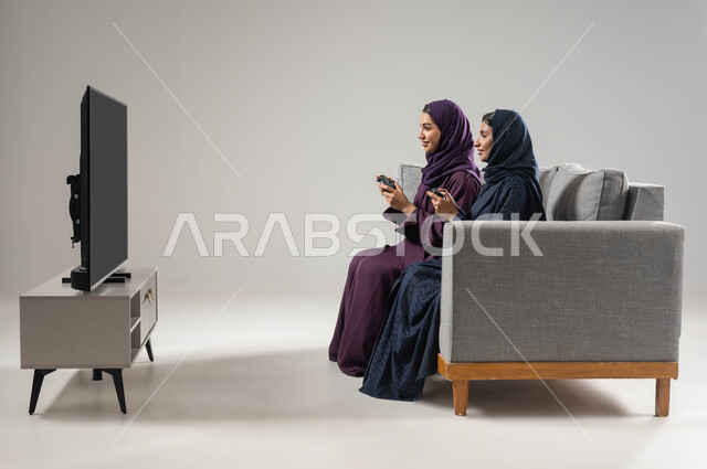 مفهوم التحدي والمنافسة، النظر بإيماءات السعادة للفوز، تقنيات حديثة مسلية, قضاء أوقات الفراغ باللعب بألعاب البلايستيشن الترفيهية، بورتريه من الجانب لإمرأتان عربيتان سعوديتان خليجيتان محجبتان تجلسان على اريكة وتحملان بيدهما عصا التحكم، صورة لكامل الجسم، خلفية رمادية