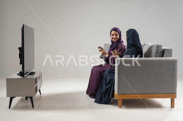 قضاء أوقات الفراغ باللعب بألعاب البلايستيشن الترفيهية، مفهوم التحدي والمنافسة، النظر بإيماءات السعادة للفوز، تقنيات حديثة مسلية, بورتريه من الجانب لإمرأتان عربيتان سعوديتان خليجيتان محجبتان تجلسان على اريكة وتحملان بيدهما عصا التحكم، صورة لكامل الجسم، خلفية رمادية