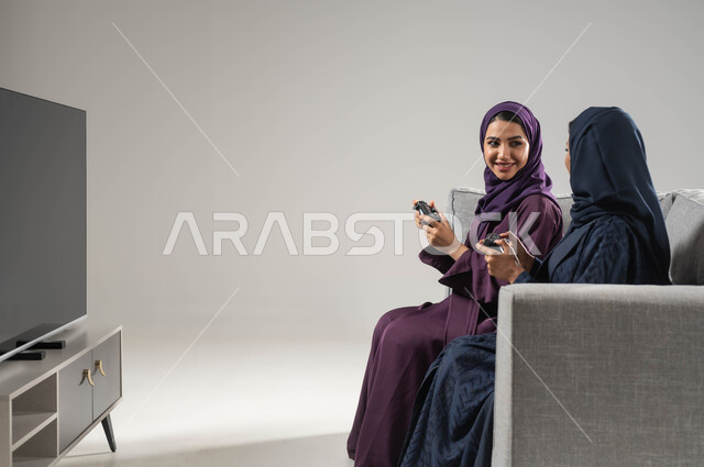 Looking with happy gestures to win, modern entertaining techniques, the concept of challenge and competition, spending free time playing entertainment PlayStation games, a portrait from the side of two veiled Saudi Arabian Gulf women sitting on a sofa and holding a joystick in their hand, gray background.
