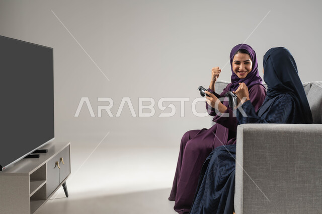 إيماءات السعادة بالفوز، تقنيات حديثة مسلية, مفهوم التحدي والمنافسة، قضاء أوقات الفراغ باللعب بألعاب البلايستيشن الترفيهية، بورتريه من الجانب لإمرأتان عربيتان سعوديتان خليجيتان محجبتان تجلسان على اريكة وتحملان بيدهما عصا التحكم، خلفية رمادية