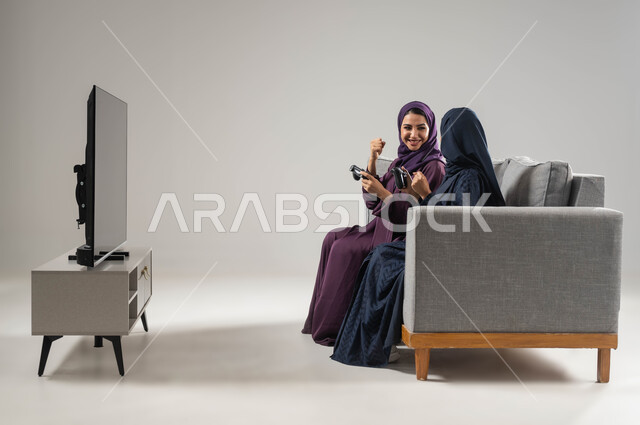 رفع اليد بإيماءات الفوز، تقنيات حديثة مسلية, مفهوم التحدي والمنافسة، قضاء أوقات الفراغ باللعب بألعاب البلايستيشن الترفيهية، بورتريه من الجانب لإمرأتان عربيتان سعوديتان خليجيتان محجبتان تجلسان على اريكة وتحملان بيدهما عصا التحكم، صورة لكامل الجسم، خلفية رمادية