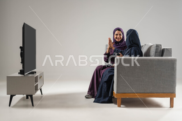 تقنيات حديثة مسلية, مفهوم التحدي والمنافسة، قضاء أوقات الفراغ باللعب بألعاب البلايستيشن الترفيهية ، بورتريه من الجانب لإمرأتان عربيتان سعوديتان خليجيتان محجبتان تجلسان على اريكة مريحة وتحملان بيدهما عصا التحكم، شاشة سوداء فارغة، صورة لكامل الجسم، خلفية رمادية