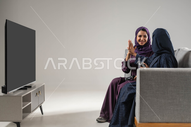 شاشة سوداء فارغة، تقنيات حديثة مسلية, مفهوم التحدي والمنافسة، قضاء أوقات الفراغ باللعب بألعاب البلايستيشن الترفيهية ، بورتريه من الجانب لإمرأتان عربيتان سعوديتان خليجيتان محجبتان تجلسان على اريكة مريحة وتحملان بيدهما عصا التحكم، صورة لكامل الجسم، خلفية رمادية