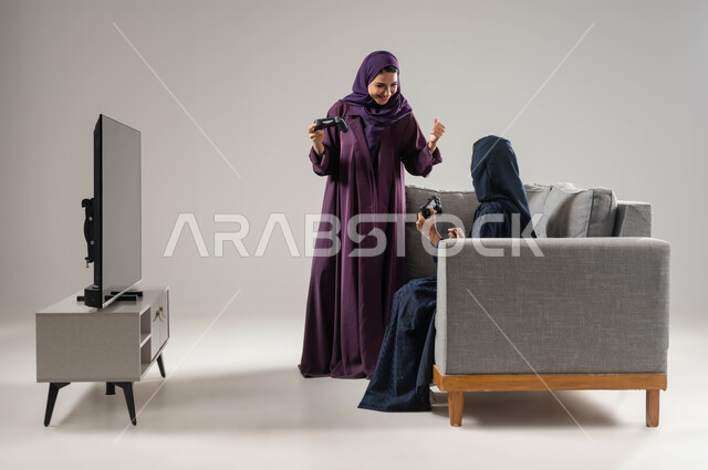 مفهوم التحدي والمنافسة، قضاء أوقات الفراغ باللعب بألعاب البلايستيشن الترفيهية ، تقنيات حديثة مسلية, بورتريه من الجانب لإمرأتان عربيتان سعوديتان خليجيتان محجبتان تجلسان على اريكة مريحة وتحملان بيدهما عصا التحكم، شاشة سوداء فارغة، صورة لكامل الجسم، خلفية رمادية