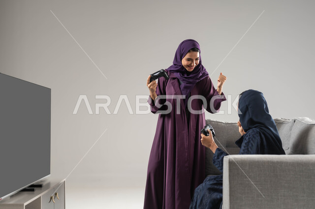 قضاء أوقات الفراغ باللعب بألعاب البلايستيشن الترفيهية ، تقنيات حديثة مسلية, بورتريه من الجانب لإمرأتان عربيتان سعوديتان خليجيتان محجبتان تجلسان على اريكة مريحة وتحملان بيدهما عصا التحكم، مفهوم التدي والمنافسة، شاشة سوداء فارغة، خلفية رمادية